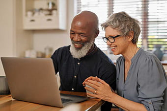 an older couple smiling and looking at their laptop together in the kitchen